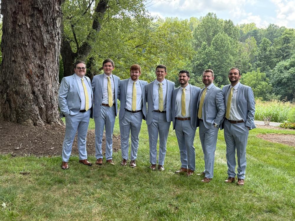 Group of men in light blue suits with yellow ties standing outdoors near a tree