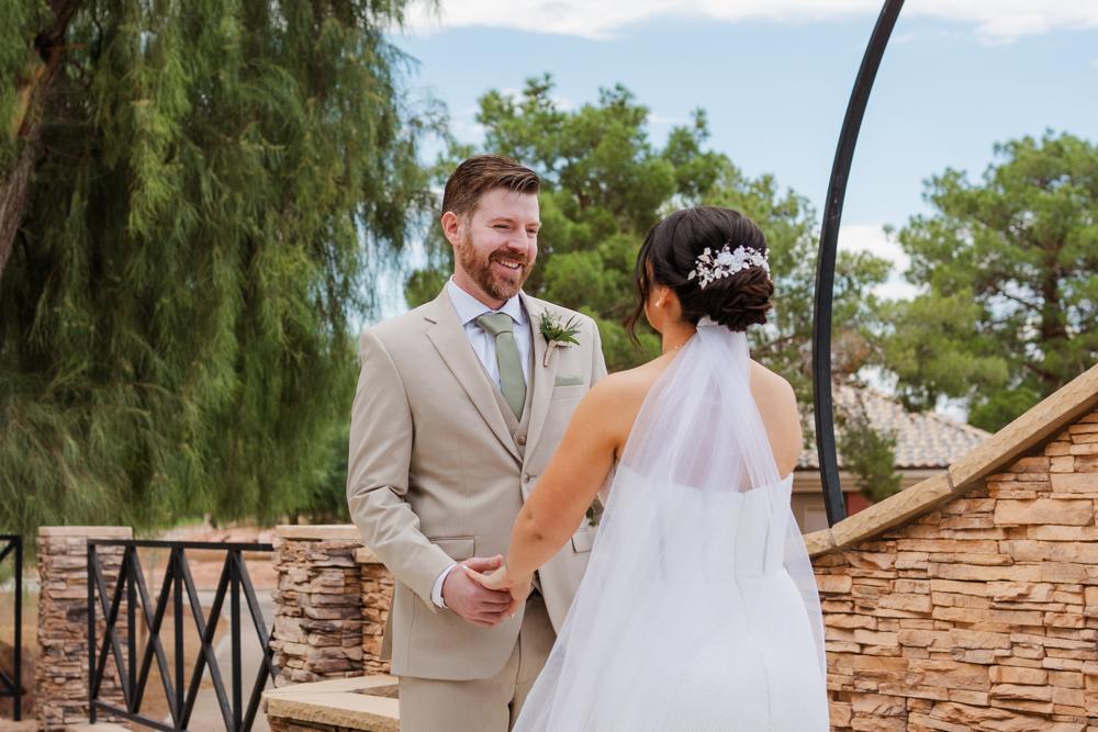 Groom in a tan suit and sage green tie holding hands with his bride outdoors.