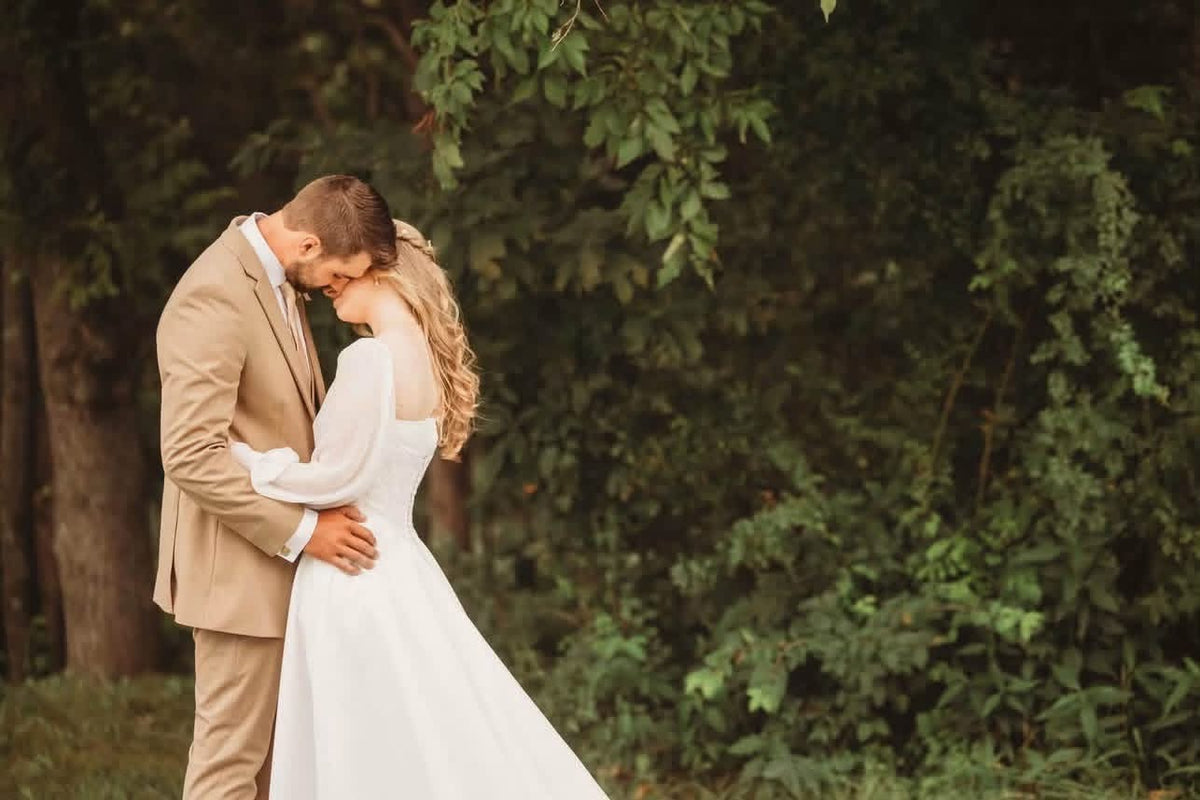 Amanda and Harlen kissing beneath leafy trees, wearing a white gown and tan suit.