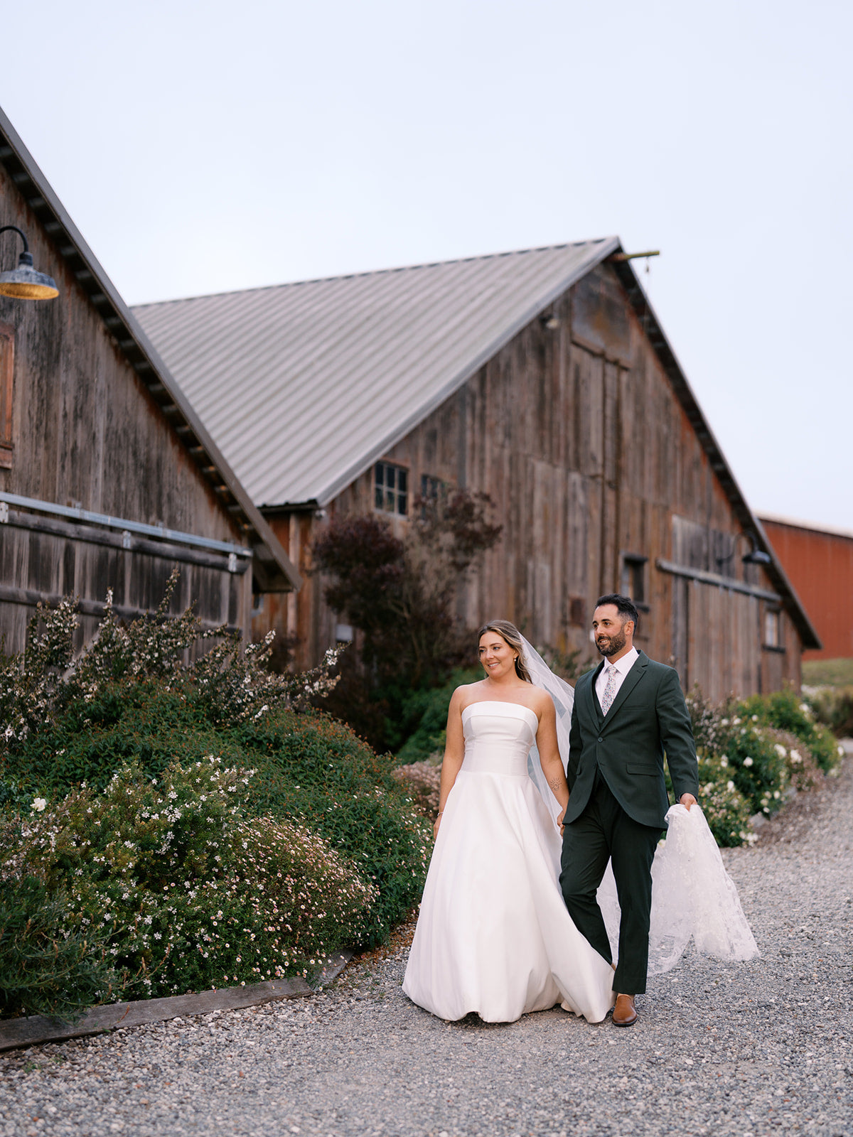 Danielle and Derick walking beside weathered wooden barns on their wedding day.