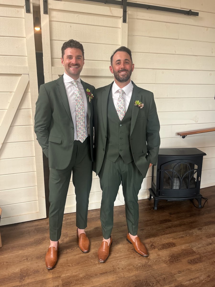 Derick and another man wearing matching olive-green suits and floral ties indoors.