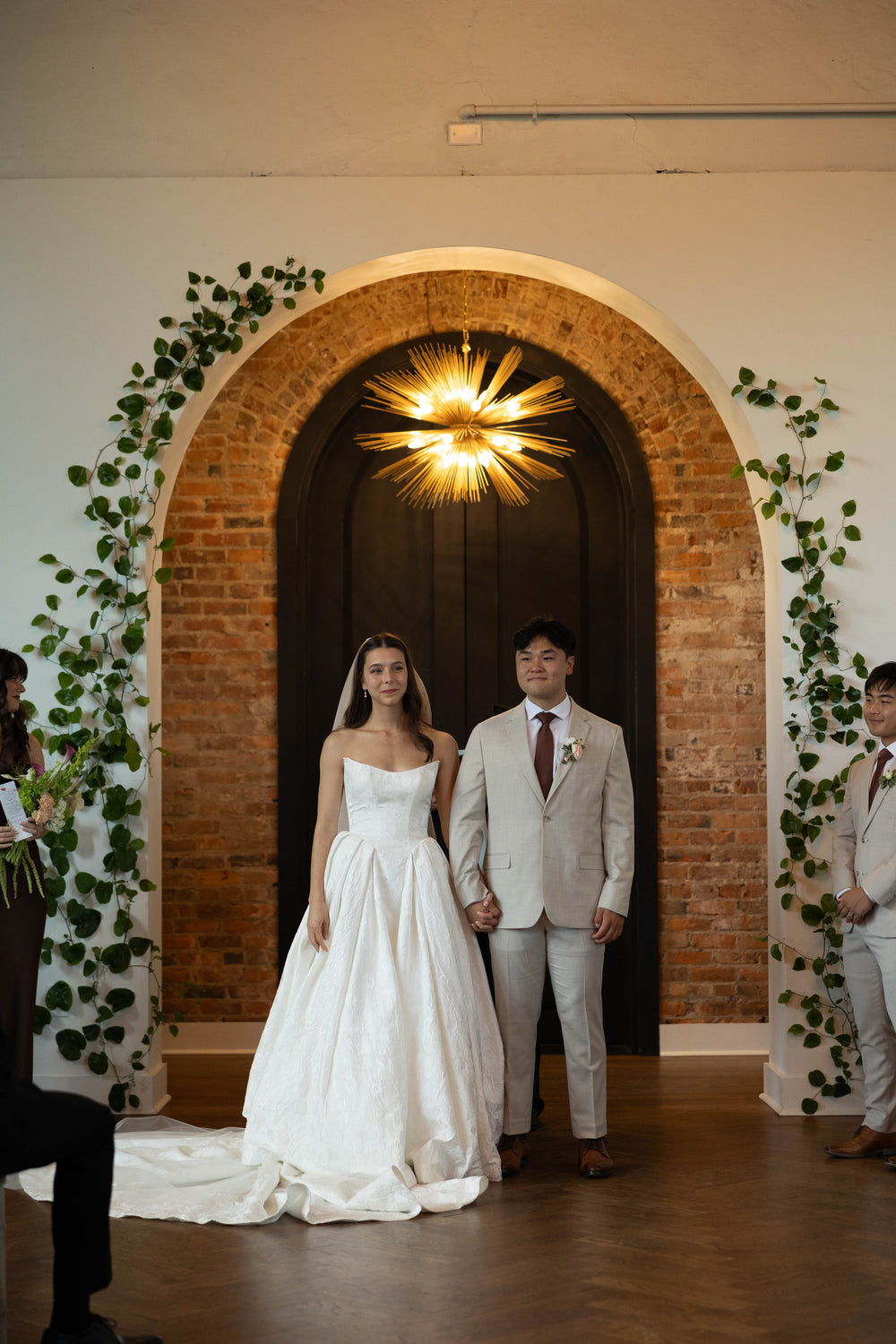 Sophia and Andrew standing together beneath a brick arch, showing his tan suit.