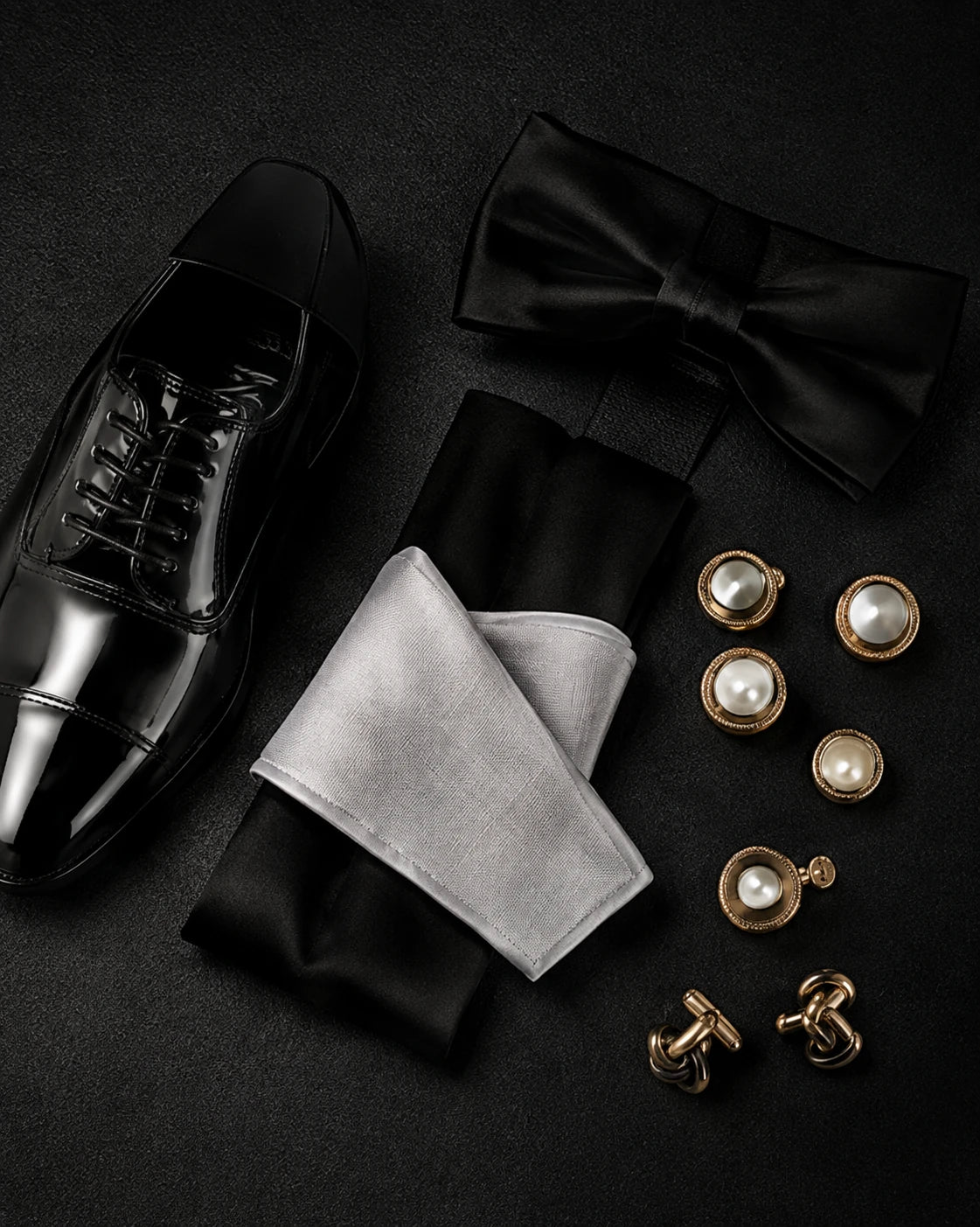 A men's formal accessories set arranged on a dark textured background, including a black dress shoe, black bow tie, black and white pocket square, and gold cufflinks and cufflinks with pearl details.