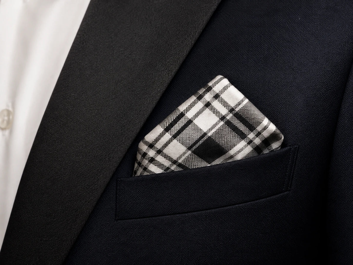 A close-up of a black plaid pocket square tucked into the breast pocket of a dark navy suit jacket, worn over a white dress shirt.