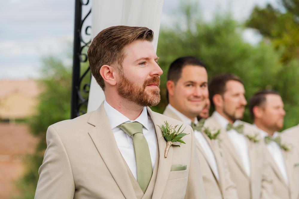 Groom in a beige suit with a green tie and boutonniere, standing outdoors with blurred groomsmen in the background.
