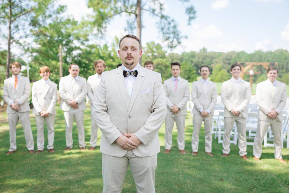 Man in a light gray suit standing in front of a group of men in similar suits outdoors.