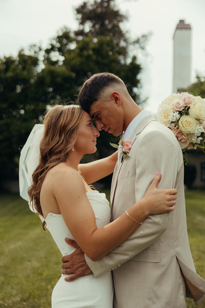 Bride embracing a groom in a light tan suit with a floral boutonniere outdoors.