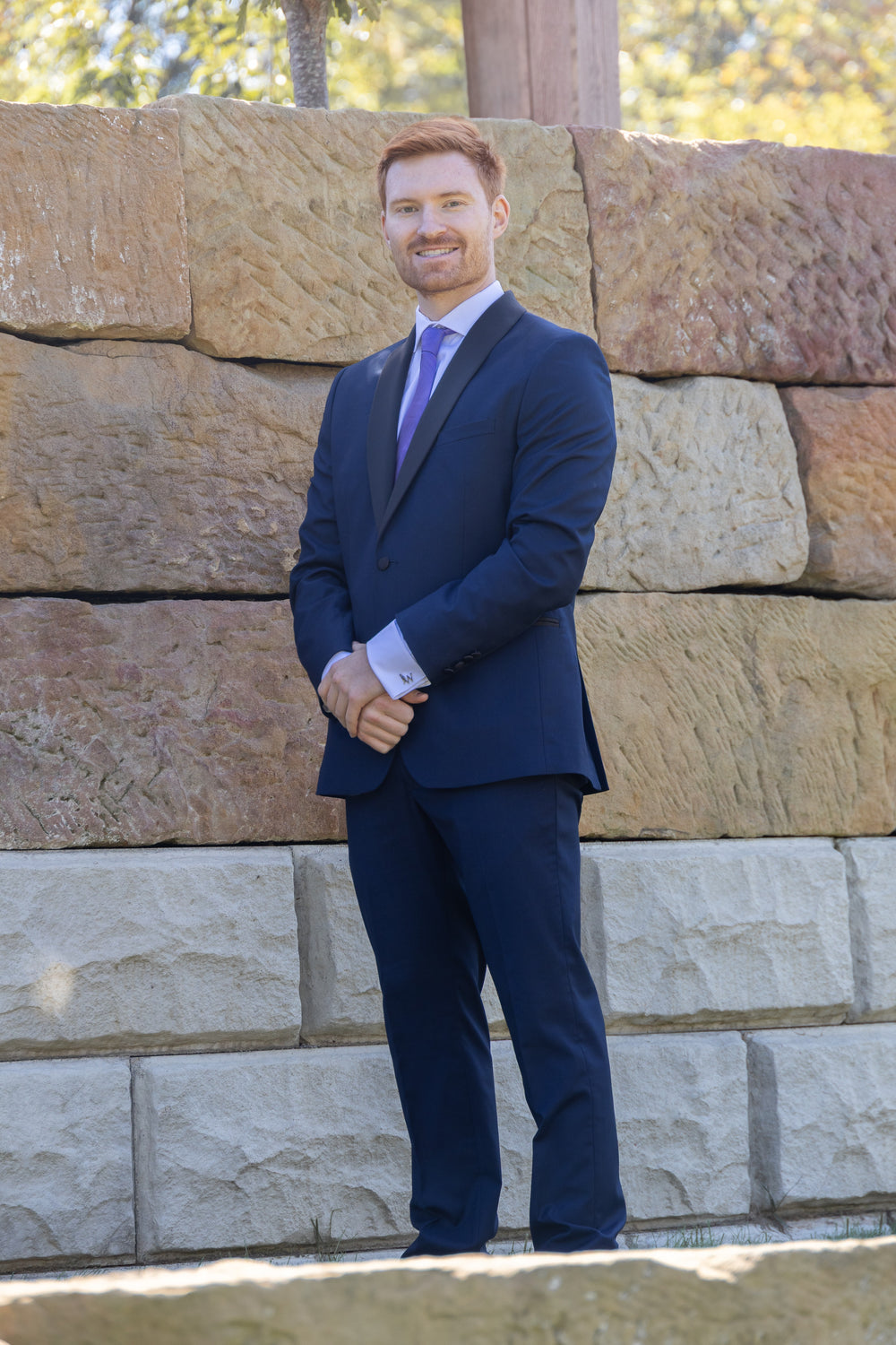 Jarrett standing by a stone wall in his navy tuxedo and purple tie.