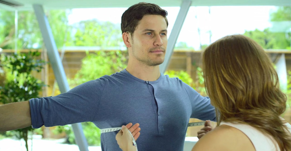 A man wearing a blue henley shirt stands indoors while a woman holds a measuring tape around his torso. The setting appears to be a bright modern interior with large windows and greenery outside.