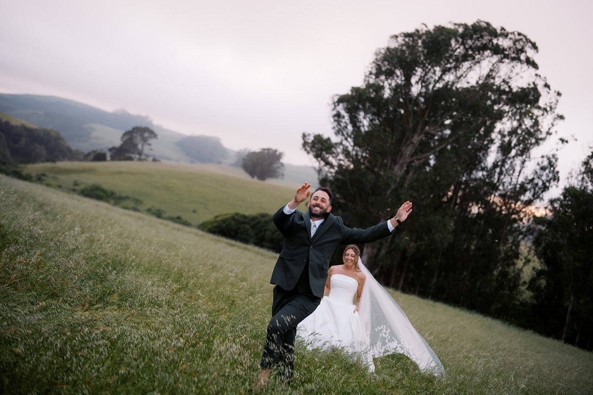 Danielle and Derick walking across a green hillside on their wedding day in Tomales, California.