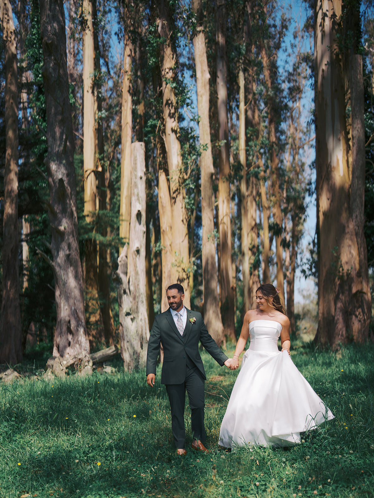 Danielle and Derick holding hands beneath tall trees, wearing a white gown and olive-green suit.
