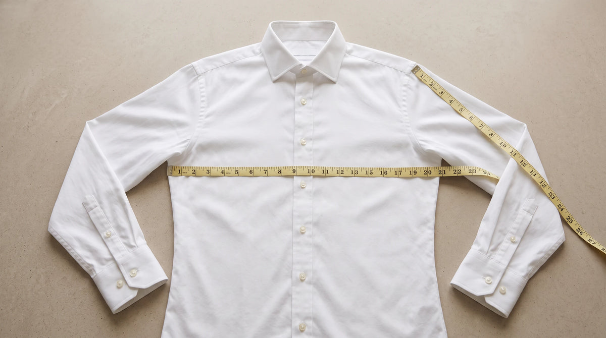 A white long-sleeve button-up dress shirt laid flat on a beige background, with a yellow measuring tape wrapped around the chest and another around the shoulder to show garment measurements.
