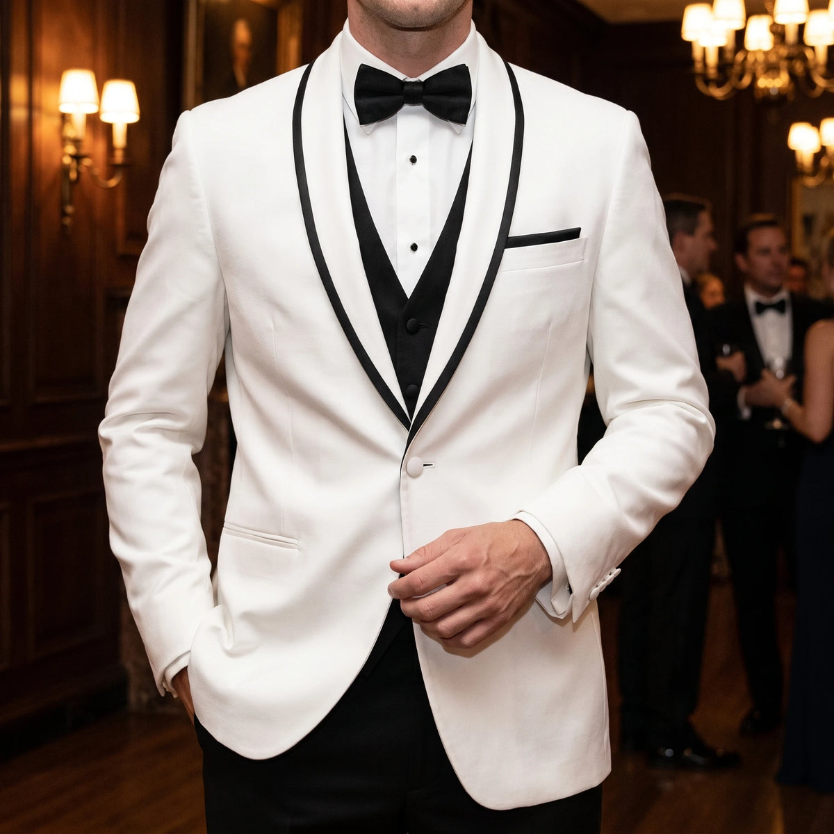 A man wearing a white tuxedo jacket with black satin peak lapels, paired with a black vest, white dress shirt, and black bow tie in a dimly lit, elegant indoor event space.