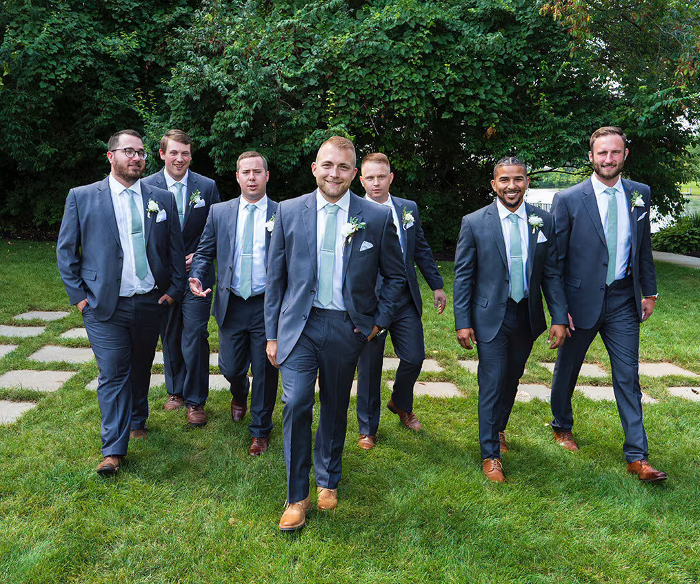 Six men in matching navy blue suits, light blue ties, and white pocket squares walking together on a green lawn with stone pavers and a backdrop of lush green trees.