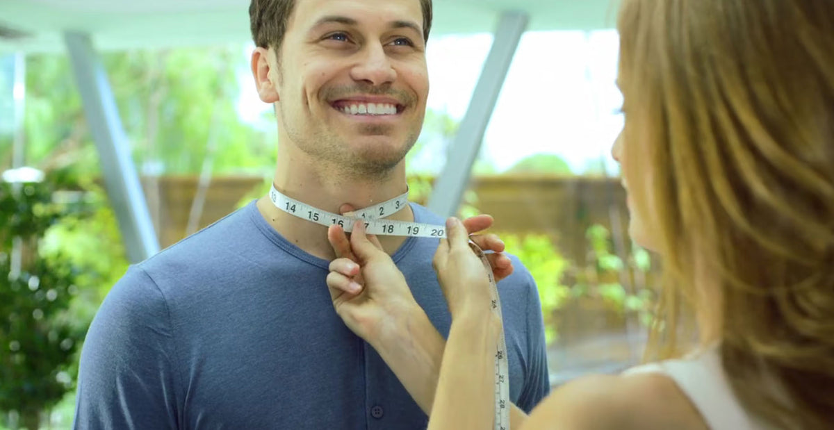 A smiling man in a blue shirt has a woman measuring his neck with a white tape measure in a bright, modern indoor setting.