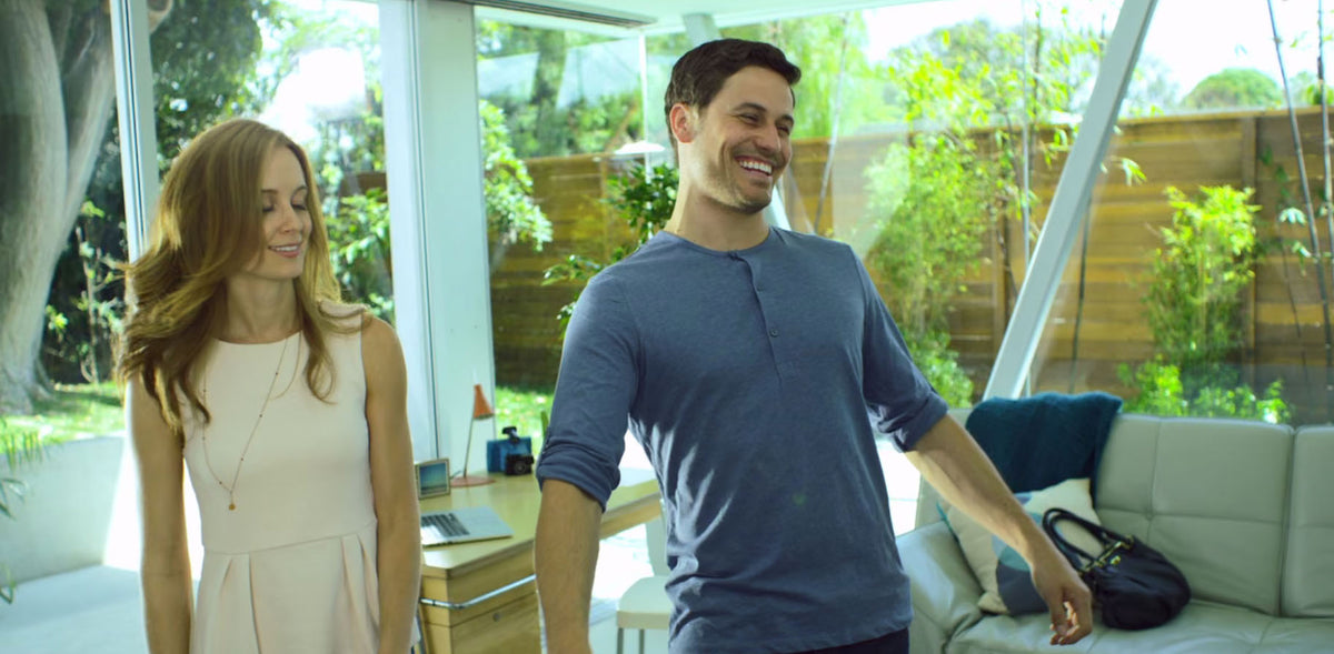 A man and a woman standing in a modern glass-walled home interior, with the man wearing a blue henley shirt and the woman wearing a light pink dress. A black handbag rests on a white sofa in the background.